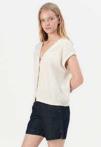 Lys beige kortermet cardigan med dyp V-hals, myk tekstur og knappelukking, paret med mørk denimshorts.