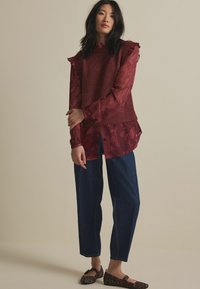 Pull en tricot sans manches bordeaux sur un chemisier transparent à motifs floraux bordeaux, associé à un pantalon en denim foncé et des ballerines à imprimé animal.