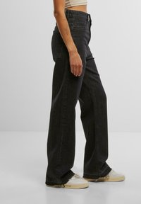 Schwarz-geschnittene Jeans mit weitem Bein und hoher Taille, versehen mit Gesäßtaschen und ausgefransten Säumen, kombiniert mit weißen Turnschuhen. Sanfte Textur.