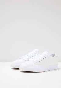 Vita canvas sneakers med gummisula, rund tå, snörning fram och logodetalj på sidan. Slät textur och minimalistisk design.