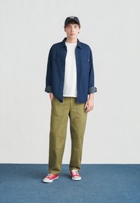 Jeune homme portant une chemise bleu marine boutonnée sur un t-shirt blanc, un pantalon vert olive, des baskets rouges et une casquette de baseball noire à rayures, debout sur un tapis bleu.