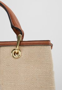Béžová taška s herringbone vzorem a hnědými koženými ušima a lemem. Obsahuje zlaté kovové doplňky s detaily smyčky a sponky.