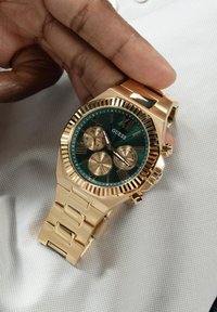 Gold watch with a green dial, featuring textured patterns, three sub-dials, and gold hour markers. Metal bracelet with a secure clasp.