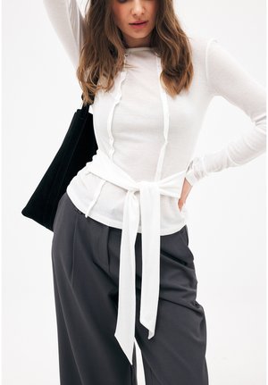 Frau trägt ein weißes langärmeliges Oberteil mit gebundenem Taillengürtel, dunkle weite Hose und trägt eine schwarze Umhängetasche.