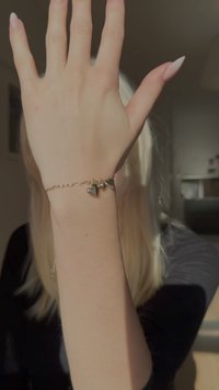 Pulseira de ouro com três pequenos pendentes em forma de coração, ligados por uma corrente delicada. A mão exibe unhas longas e de cor clara.