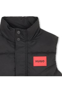 Μαύρο αμάνικο μπουφάν με διπλωμένο γιακά, με δύο μαύρα κουμπιά και μια κόκκινη ορθογώνια ετικέτα με την επιγραφή "HUGO" σε μαύρο κείμενο.