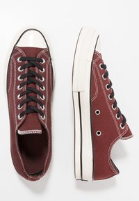Zapatillas de lona color marrón con cordones negros, puntera de goma blanca y suela de goma. Presenta costuras blancas y ojales de metal.