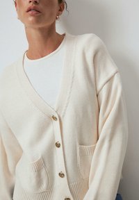Crème-kleurige cardigan met een diepe v-hals, gouden knopen en voorzakken. De stof heeft een zachte textuur met ribgebreide accenten.