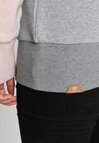 Sudadera gris y rosa con dobladillo acanalado, textura suave y parche de logo de cuero marrón. Cuenta con mangas largas y un ajuste casual.