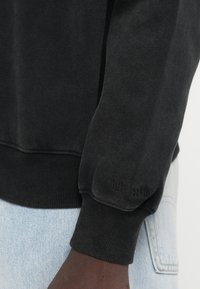 Černá mikina s kartáčovanou texturou, žebrovanými manžetami a lemem. Diskrétní logo značky vyražené na rukávu. V kombinaci se světle modrým denimem.