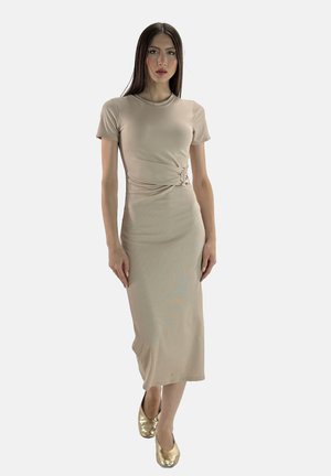 Modelo con un vestido beige ajustado y un cinturón lateral, de pie con confianza. Lleva zapatos dorados, contra un fondo blanco liso.