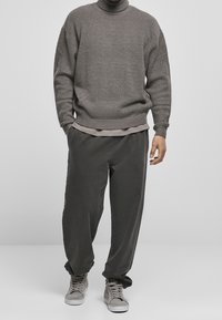 Mann in grauem Rollkragenpullover, lockerer dunkelgrauer Hose und grauen High-Top-Sneakern steht mit entspannten Händen.