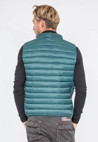 Gilet matelassé teal avec un col montant, présentant des boudins horizontaux. Porté par-dessus une chemise noire à manches longues, mettant en valeur un tissu texturé.
