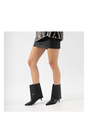 Vrouw die zwarte kittenhakken draagt met puntige neus en een zwarte leren minirok met een zebrapatroon top, tegen een witte achtergrond.