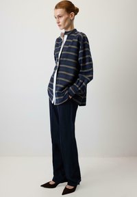 Skjorte med knapper i marineblå med vandrette guldstriber, kombineret med marineblå bukser med brede ben og spidse brune flade sko. Glat stof og struktureret design.