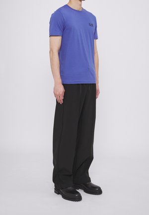 Uomo con maglietta blu a maniche corte, pantaloni neri a gamba larga e scarpe nere, in piedi davanti a uno sfondo bianco uniforme.