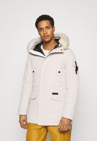Parka di colore chiaro con cappuccio foderato in pelliccia, chiusura a zip e bottoni a pressione, due tasche frontali e una tasca laterale con hardware regolabile.
