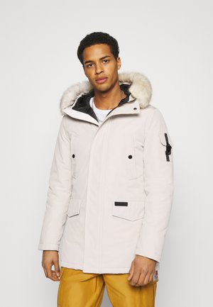 Parka di colore chiaro con cappuccio foderato in pelliccia, chiusura a zip e bottoni a pressione, due tasche frontali e una tasca laterale con hardware regolabile.