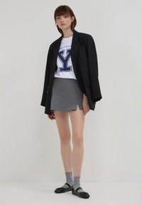 Giovane donna che indossa un blazer nero sopra una t-shirt bianca con logo dell'Università di Yale, una mini gonna grigia, calzini grigi e ballerine nere, in piedi contro un muro bianco.