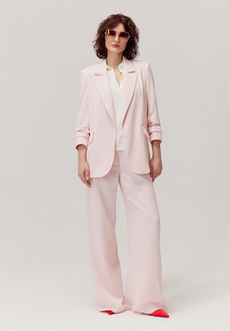 Różowa marynarka i szerokie spodnie w lekkim materiale. Marynarka ma klapy i delikatne wykończenia przy mankietach. Noszona z białą bluzką i czerwonymi akcentami na butach.