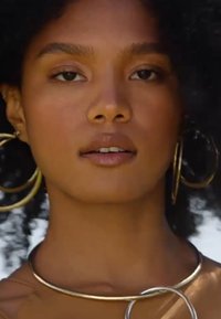 Des boucles d'oreilles en or et un collier en métal mettent en valeur une peau lisse et des cheveux bouclés, sur un fond neutre.