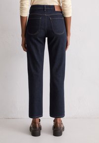 Donkerblauwe rechte jeans met een hoge taille, leren label achterop en subtiele gouden stiksels. Draagt bij donkere bordeaux platform schoenen.