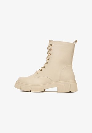 Beżowe botki za kostkę wykonane z fakturowanej skóry, z okrągłym noskiem, grubą podeszwą, złotymi oczkami i pętelką z tyłu ułatwiającą zakładanie.