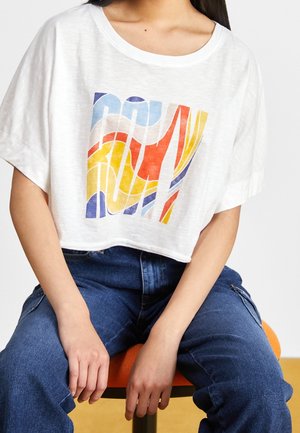Kobieta siedzi na pomarańczowym krześle, ubrana w biały krótki T-shirt z wielokolorowym abstrakcyjnym wzorem i niebieskie dżinsy.