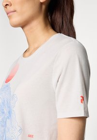 Ljust grå bomullströja med en blå linjegrafik och röda detaljer. Korta ärmar med en liten logotyp på vänster ärm.