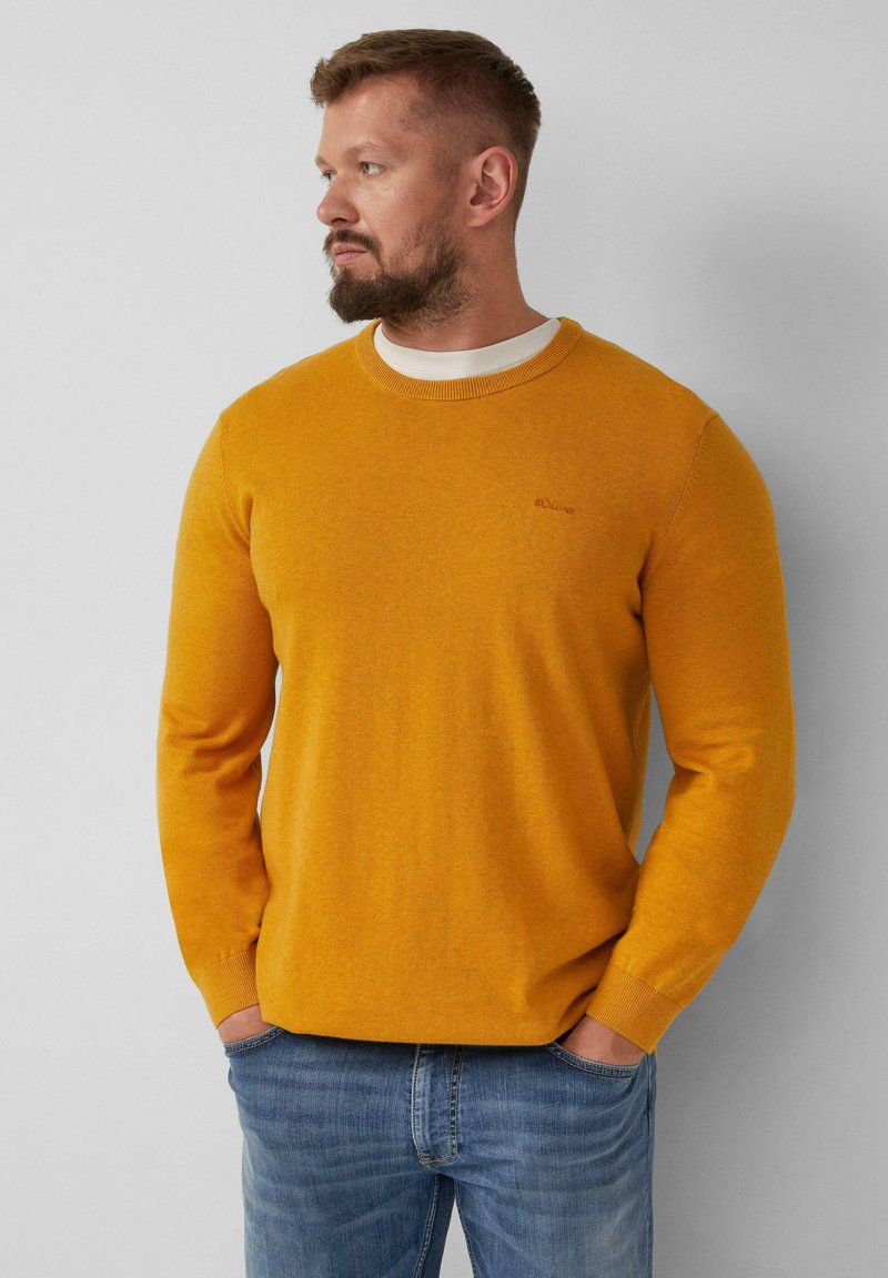 Mann mit Bart, der einen senfgelben Pullover über einem weißen Hemd und blauen Jeans trägt, steht mit Händen in den Taschen und schaut nach links.
