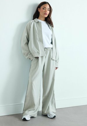 Kobieta stojąca w pomieszczeniu, ubrana w luźne, paskowane jasnoszare spodnie, pasującą kurtkę na zamek, białą koszulkę i białe trampki.