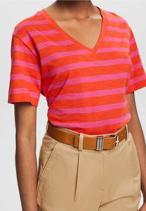 Person trägt ein rot-rosa gestreiftes V-Ausschnitt-T-Shirt, das in beige Hosen mit einem hellbraunen Gürtel mit silberner Schnalle gesteckt ist.