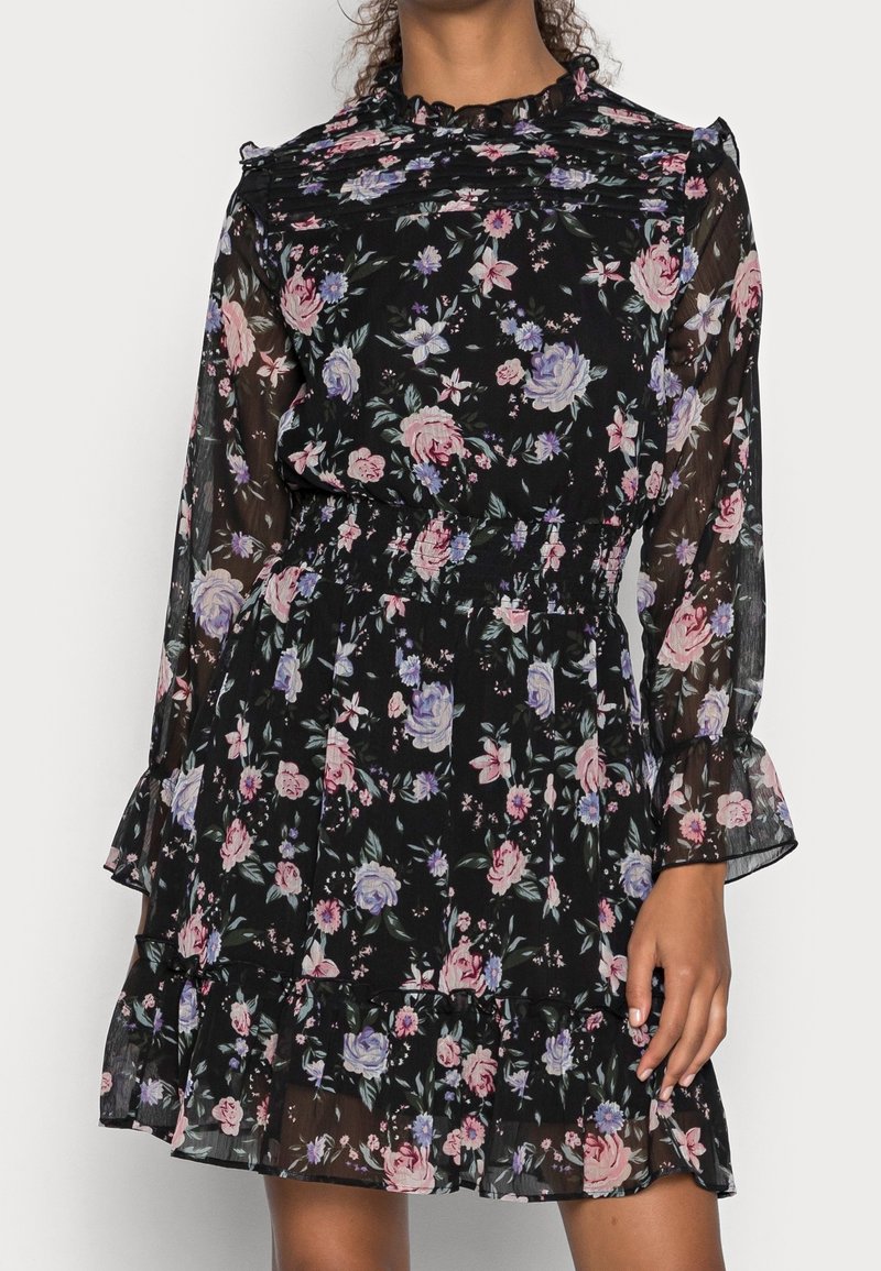 Robe noire transparente à fleurs avec manches longues, col à volants, taille cintrée et jupe à volants ornée de fleurs roses et violettes.