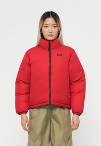 Chaqueta de plumas roja con cuello alto, cremallera negra y logotipo. Presenta una textura acolchada y puños elásticos. Combinada con pantalones caqui.