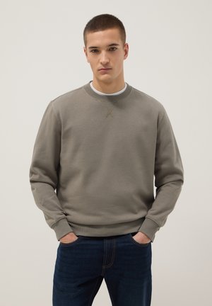 Jonge man met kort haar, gekleed in een taupekleurige trui met ronde hals en donkerblauwe spijkerbroek, staand met handen in de zakken tegen een effen achtergrond.