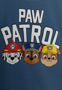 Blå bomullströja med texten "PAW PATROL". Grafiken visar tre tecknade hundar i hattar: brandman, polis och byggarbetare.