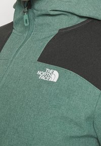 Grön och svart zip-up jacka med texturerad tyg, hög krage och en vit logotyp på bröstet. Kontrastpanel på axeln.