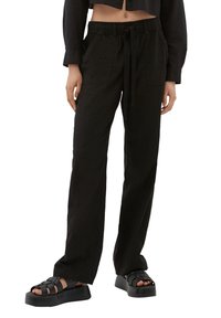 Pantalon noir à coupe ample avec taille élastique, cordon de serrage et poches latérales, accompagné de sandales noires épaisses. Tissu lisse et léger.