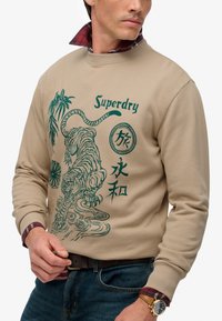 Felpa beige con grafica di tigre verde, bambù, caratteri giapponesi e testo "Superdry" indossata sopra una camicia scozzese rossa e jeans blu.
