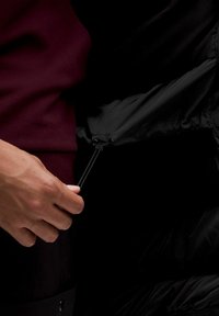 Hand die het binnenste trekkoord van een zwarte jas aanpast, met een donker bordeauxkleurig shirt, close-up weergave die zich richt op de hand en het aanpassingsonderdeel.
