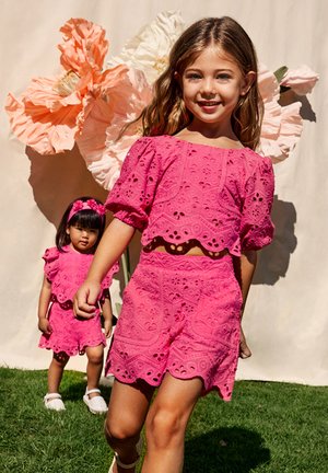 Due bambini indossano abiti in tessuto eyelet rosa brillante abbinati con bordi scallopati. Il bambino più grande ha un top corto e pantaloni shorts; il più piccolo indossa un vestito.