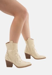 Botas de vaquero beige con un acabado texturizado, patrones de costura en relieve y acentos de tachuelas, que cuentan con un tacón cuadrado y cremallera lateral.