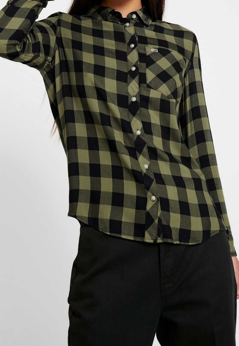 Personne portant une chemise à manches longues à carreaux verts et noirs boutonnée, associée à un pantalon noir, sur un fond clair uni.