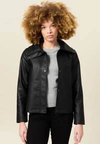 Giacca di pelle nera con un ampio colletto, bottoni a pressione e zip sul davanti. Indossata sopra un maglione grigio, su uno sfondo neutro.