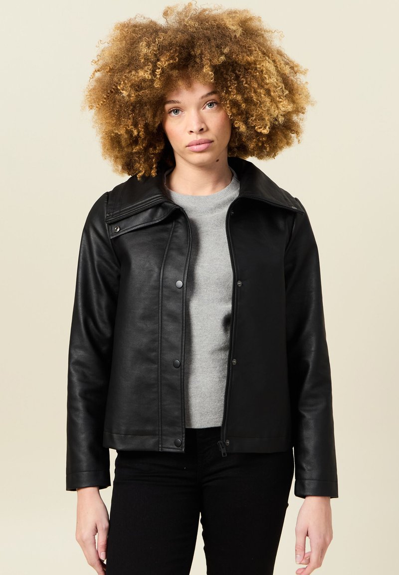Giacca di pelle nera con un ampio colletto, bottoni a pressione e zip sul davanti. Indossata sopra un maglione grigio, su uno sfondo neutro.