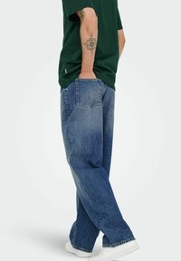 Mörkgrön t-shirt med rund halsringning, löst sittande urtvättade blå denimjeans med rak passform, vita sneakers och synliga bakfickor.