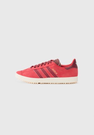 Röda mocka sneakers med tre mörkröda ränder, rund tå, beige gummisula och en gyllene "Gazelle"-logotyp på sidan.