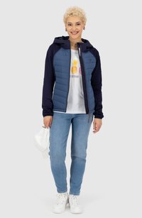 Marineblau gepolsterte Jacke mit Kapuze über einem weißen Graphic-T-Shirt, kombiniert mit hellblauen Jeans und weißen Sneakers, hält eine weiße Kappe.