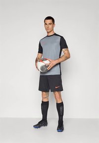 Conjunto de ropa deportiva Nike que incluye una camiseta de manga corta gris y negra, pantalones cortos negros y espinilleras con un balón de fútbol con patrón.