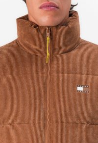Bruine corduroy jack met ritssluiting en gele ritssluitingtrekker, voorzien van een klein Tommy Jeans-logo op de borst, gedragen door een persoon van hals tot onder.